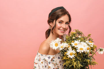 Close-up well-groomed young caucasian girl smiling with teeth on pink background. Brown-haired woman with gathered hair, dressed in blouse with open shoulders, holds bouquet of wildflowers in hands.