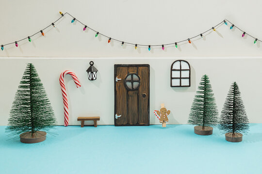 Mini elf or mouse house diy toy gift for kids made with tiny door, window with lantern and Christmas green tree of pipe brushes. Little bulb garland on white wall. Holiday decor on blue background.