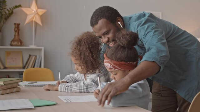 Medium shot of happy African-American man helping his two cute daughters with school homework studying together at cozy home