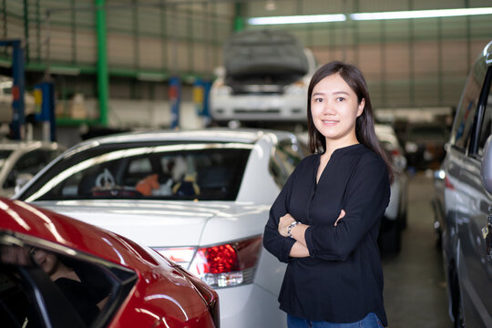 Portrait of beautiful Asian woman posing for photograph beside the car in garage, woman bring her vehicle to repair in garage. Satified Asian young woman standing beside the car in garage.