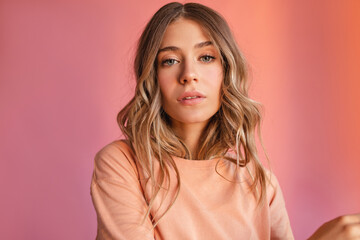 Obraz premium Close-up of calm caucasian young woman looking at camera on pink background. Blonde with wavy, loose hair wears peach-colored T-shirt. Female beauty concept