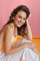 Naklejka premium Lovely caucasian young girl with pleasant appearance sits sideways on floor in room with pink wall. On colored background, cute smiling brown-haired woman in summer light clothes.