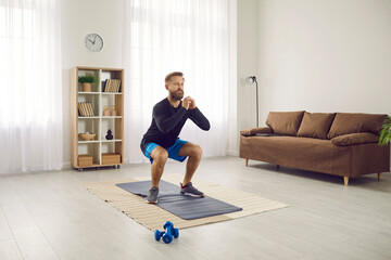 Portrait of active focused millennial man doing morning sports at home to keep fit. Athletic fit man in sports uniform performs squat exercise in his spacious bright apartment with minimalist interior