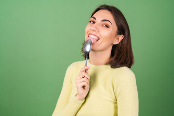 Obraz premium Young woman on a green background in a yellow sweater with appetite eats from a tablespoon, enjoying