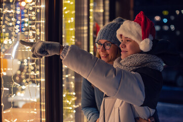 Pretty Woman and her son in santas hat looking and dreaming in illuminated shop window. Xmas presents holidays, or shopping on New Year or Christmas concept