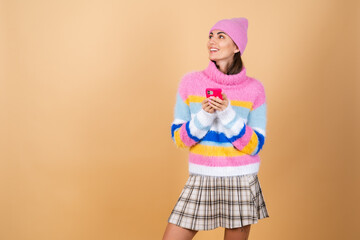 Young woman on a beige background in a knitted sweater and a hat with a mobile phone laughing with surprise and fun, reading messages, jokes, congratulations