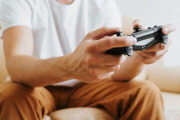 Obraz na płótnie Canvas Close-up of gamer's male hands holding wireless game controller with joysticks. Side view of man fun, playing video game on console while sitting on couch at home. Cropped, selective focus on front