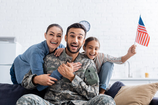 Positive family with american flag looking at camera near man in military uniform at home