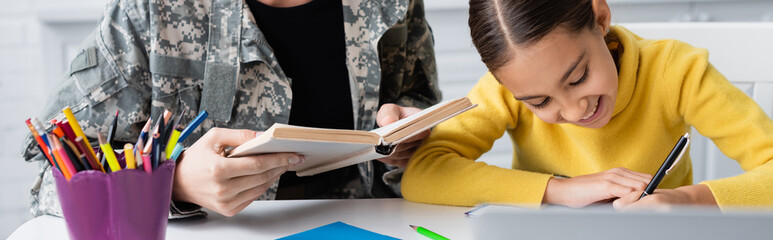 Smiling kid holding pen near mother in military uniform with book near laptop at home, banner