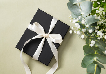 a black gift box with a white ribbon on a green background with a bouquet of eucalyptus and gypsophila. The concept of a holiday or gift card. Top  view and copy space
