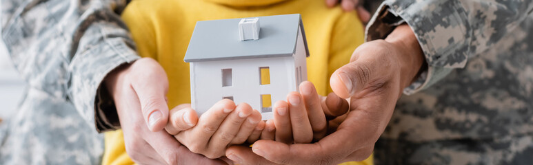 Cropped view of parents in military uniform holding model of house with daughter, banner - Powered by Adobe