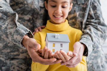 Model of house on hands of blurred kid near parents in military uniform at home