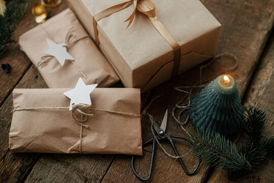 Stylish christmas gifts wrapped in craft paper, candle, scissors, fir branches and bells on rustic wood. Stylish simple scandinavian xmas presents, atmospheric moody image. Eco friendly holidays