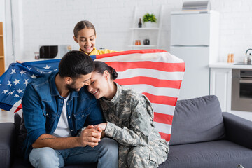 Smiling preteen girl holding american flag near mother in military uniform and father on couch
