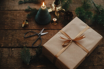 Stylish christmas gift wrapped in craft paper, candle, scissors, fir branches on rustic wood. Modern simple eco friendly xmas present, scandinavian atmospheric moody image. Happy Holidays!