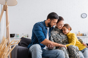 Positive woman in military uniform sitting near child and husband on couch at home