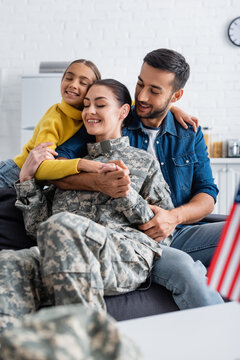 Smiling kid embracing mother in military uniform near father and blurred american flag at home