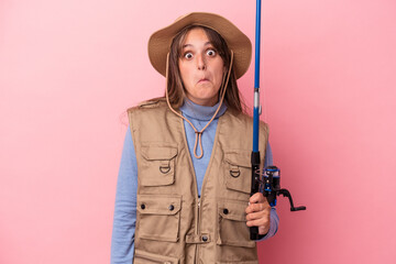 Young caucasian fisherwoman holding a rod isolated on pink background shrugs shoulders and open eyes confused.