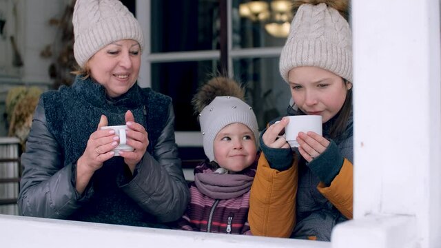 Ederly woman, a loving grandmother with two granddaughters, drink tea near her house in winter, a mature woman takes care of her little granddaughter, gives her a sip of warm tea. Beautiful portrait