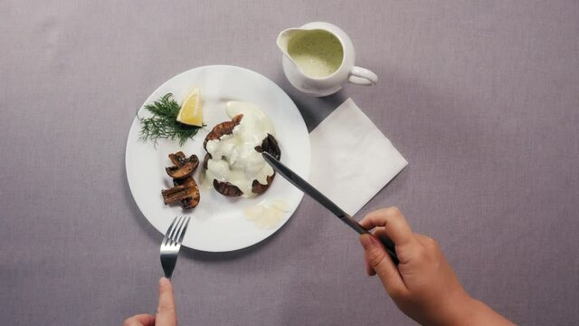 Chef hands hold fork and knife above white plate served with cooked champignon mushrooms with lemon greens and sauce closeup