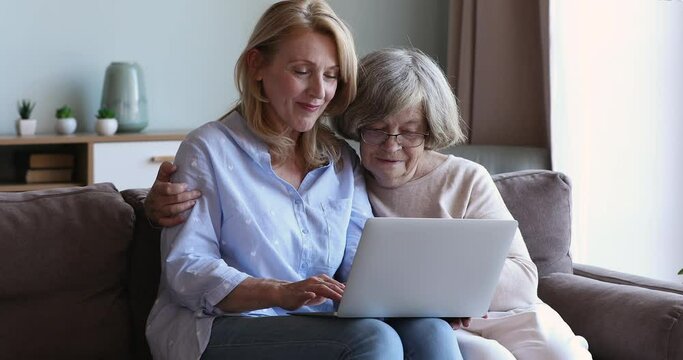 Interested senior mother pensioner embrace adult daughter sitting on sofa engaged in web shopping using laptop. Elderly lady learn to pay utility bills online via pc with help of younger female friend