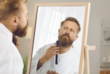 Obraz premium Reflection in mirror of handsome man combing his beard in preparation for work or important event. Serious Caucasian man in white bathrobe takes care of his beard in morning. Concept of male beauty.