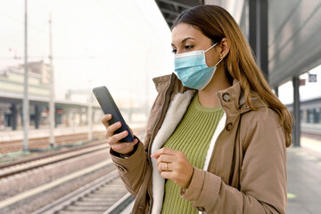 Portrait of beautiful woman with surgical mask using her smart phone waiting her train at station...