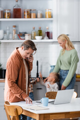 freelancer talking on mobile phone while working with documents in kitchen near laptop and blurred family