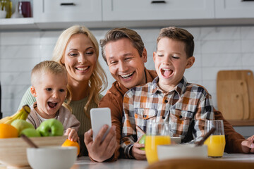cheerful man taking selfie on mobile phone with wife and sons during breakfast in kitchen