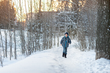 A cheerful and handsome boy in blue clothes runs along a snow-covered path in the park. Winter activities.