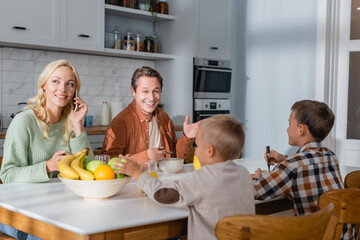 cheerful man talking to sons during breakfast while wife calling on mobile phone