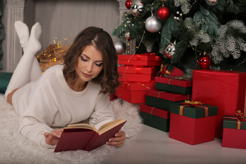 a young beautiful girl in a white tunic and knee socks is lying on a rug by the fireplace with a book in her hands next to a Christmas tree gift boxes