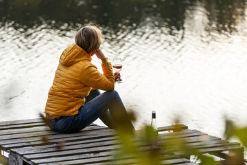 Woman in a yellow jacket relaxing on a wooden pier on a lake with a glass of rose wine watching fall sunset alone.