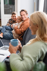 blurred woman with tea cup working on laptop near happy husband with sons on couch