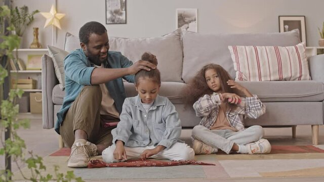 Slowmo shot of African-American man spending time with two lovely daughters at home, chatting while getting their hair done