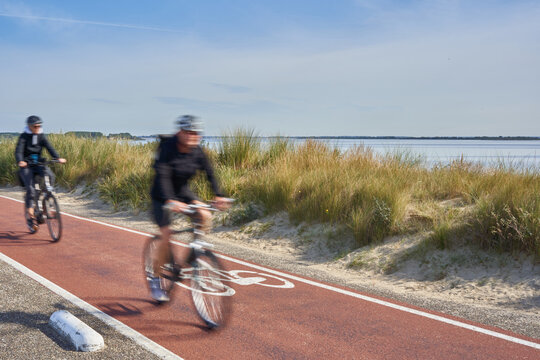 Bicycle path in the Netherlands. Dune with grass in front of a blue sea.1 pair of bicyclists motion blurred. Blue sky for text free space. In South Holland on the Brouwersdam at the Grevelinger Sea.
