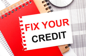 On a light background - reports, a magnifying glass, brown and red notepads, and a white notepad with the text FIX YOUR CREDIT. Business concept