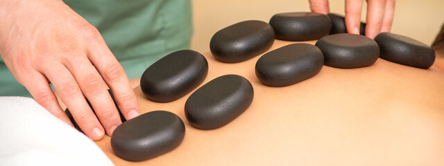 Young caucasian woman receiving back massage with black stones by masseur in spa salon. A woman getting a spa treatment.