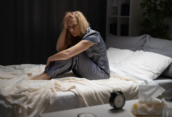 Pensive mature woman holding hands on her head while sitting on bed at night. Caucasian lady in...