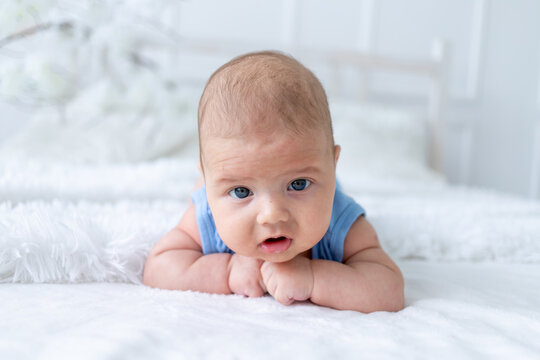 cute baby boy three months old in a blue bodysuit lies on his stomach on a white bed at home, the newborn learns to hold his head