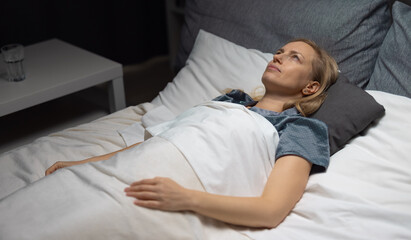 Pensive mature woman in pajamas lying awake in bed with white linen and looking at ceiling....