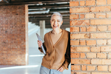 Short hair blond woman designer with laptop in loft empty space. Portrait of middle age smiling woman looking at camera. Successful businesswoman y standing in creative future office.