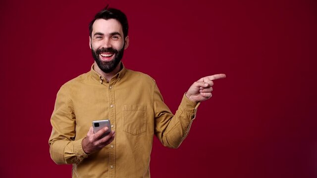 the guy holds the phone in his hands and points to the side, brand, logo, product, empty space, he smiles, is dressed in a brown shirt, the background is red, bearded, black-haired