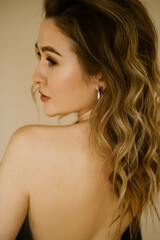 portrait of young beauty caucasian woman with her bare back covered by a jacket , a girl with makeup and curly hair , a dark eyes lady with earrings-rings and bright makeup on light background