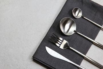 Knife, spoons and fork with dark textile napkin on grey stone background. Table setting. Top view. 