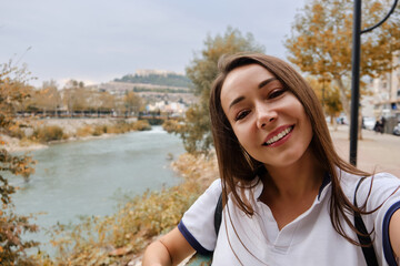 Portrait of young adult caucasian woman. Smiling brown-haired woman takes a selfie opposite autumn landscape with river at background