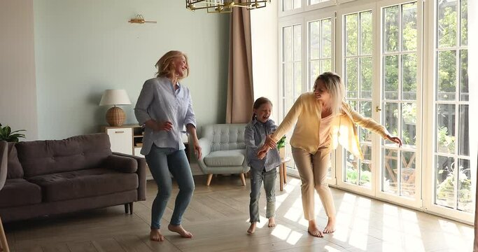 Overjoyed 3 female generations dance jump barefoot on warm wooden floor at modern living room near panoramic window. Active aged granny young mom junior school age daughter have fun at cozy apartment