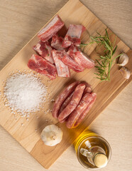 Pork ribs, sausage segments, rock salt, garlic, onion, rosemary and olive oil on a light rustic wood on a table, top view.