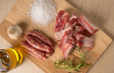 Pork ribs, sausage segments, rock salt, garlic, onion, rosemary and olive oil on a light rustic wood on a table, top view.