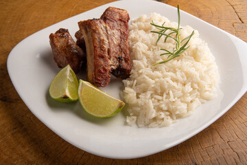 Fried pork ribs, rice and condiments on a white plate arranged on light rustic wood, selective focus.